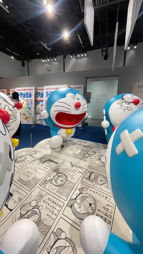 Life-size statues on the manga-page floor, blue carpet, ceiling lights.